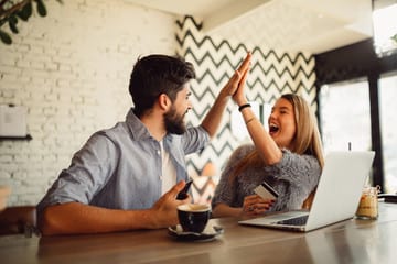 Two happy coworkers celebrating success with a high five in a cafe, with a laptop and coffee on the table