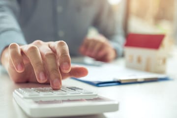 Close-up of a person's hand using a calculator with a model house and clipboard in the background, symbolizing real estate and financial planning.