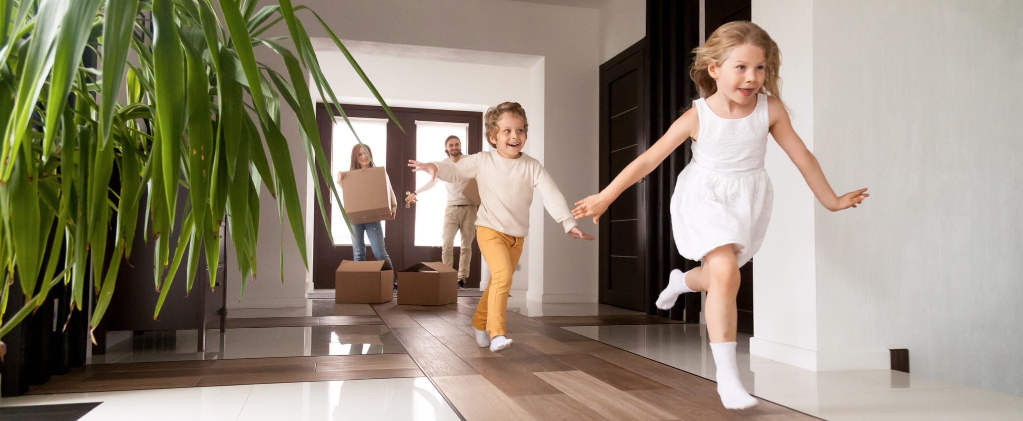 Children running excitedly through their new home with parents carrying moving boxes in the background.