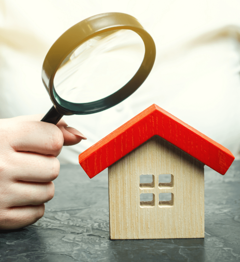 Person holding a magnifying glass over a small wooden house model with a red roof